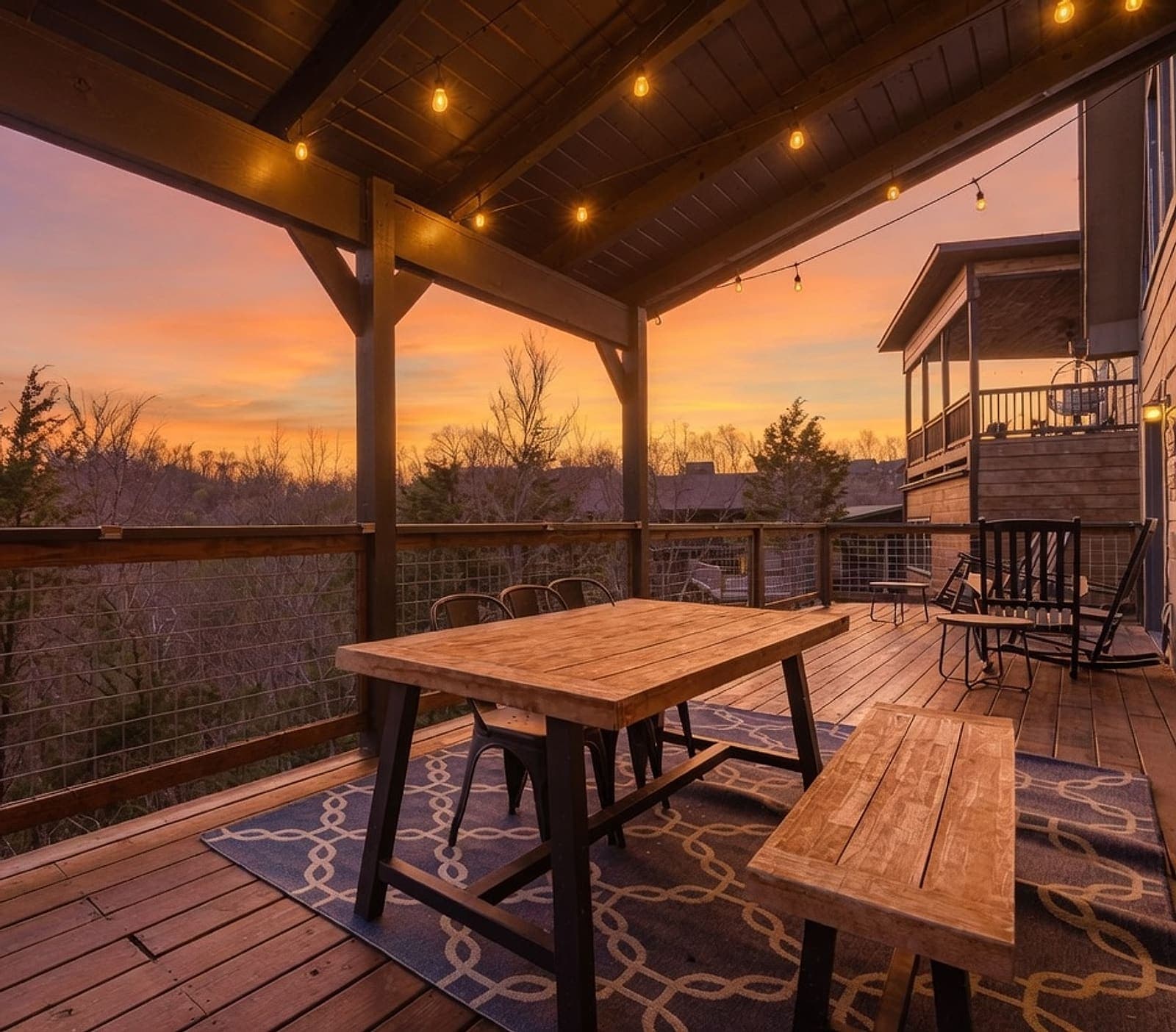Sunset from the lodge’s covered deck — string lights glowing over the farmhouse table, the Smokies’ tree line against a pastel sky
